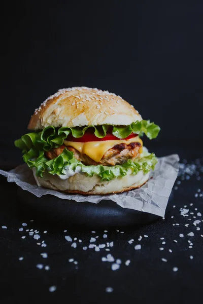 Appetizing fresh homemade burger, with chicken cutlet, lettuce, tomatoes, cheese and sauce. On a wooden board on a black background