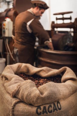 Jute bag full of cocoa beans in a chocolate maker workshop, with a male chocolatier working on conching and melanger equipment on the background clipart
