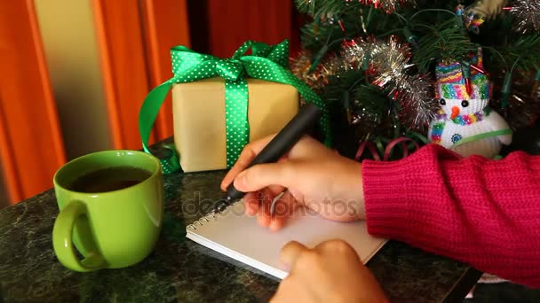 Mujer escribiendo lista de deseos, con regalos y árbol de Navidad en el fondo — Vídeos de Stock
