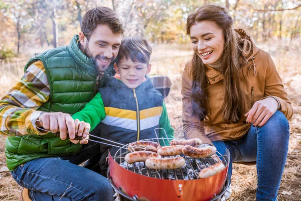 Веселі батьки з сином смажать ковбаски — стокове фото