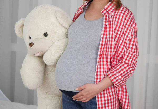 a pregnant woman dressed in a gray t-shirt and a bright shirt and holds a large toy bear in her right hand, and holds her stomach with her left hand