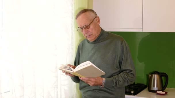 Пожилой мужчина читает книгу, стоя у окна — стоковое видео