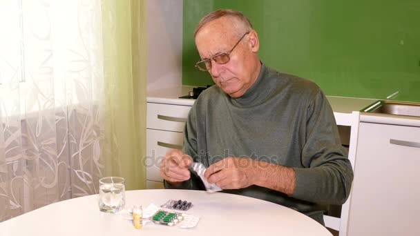 Starý muž sedí u stolu a při pohledu na prášky — Stock video