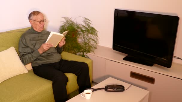 Viejo sentado en el sofá y leyendo un libro en la sala de estar — Vídeos de Stock