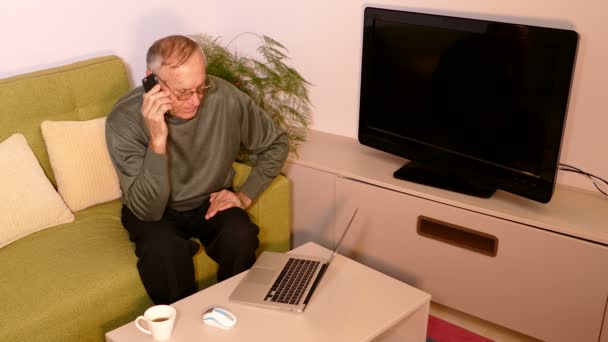 Gammal man sitter i vardagsrummet prata telefon och använder bärbar dator — Stockvideo