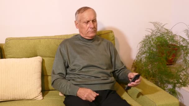 Старик сидит на диване и включает телевизор с пультом дистанционного управления — стоковое видео
