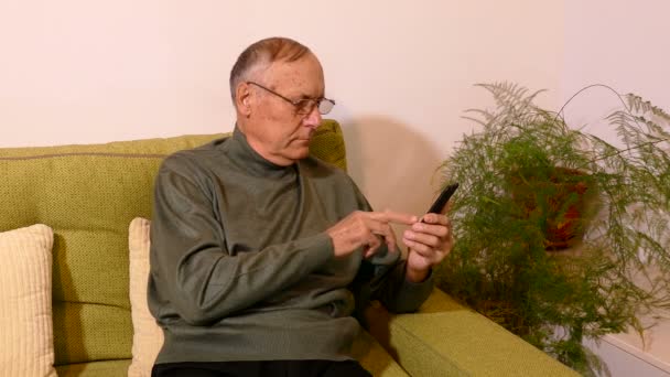 बूढ़ा आदमी लिविंग रूम में सोफा पर बैठे और अपने सेलफोन का उपयोग करते हुए — स्टॉक वीडियो