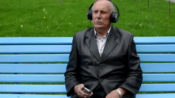 Viejo escuchando música sentado en el banco en el parque — Vídeo de stock