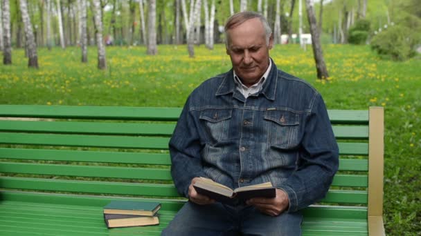 Gamling sitter på benken i parken og leser en bok. – stockvideo