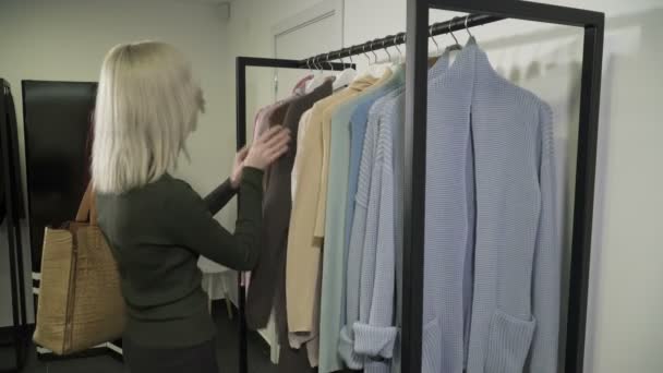 Zbliżenie, młoda kobieta blonde, zbierając ubrania w butiku — Wideo stockowe