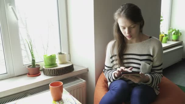 Nastolatek dziewczyna siedzi na fotelu i plaing z tabletu podczas picia herbaty — Wideo stockowe