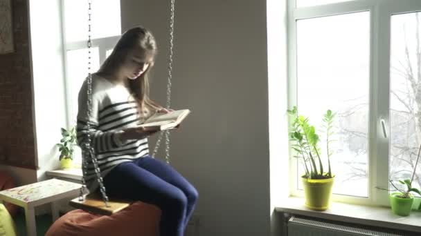 Tiener meisje zittend op een swind in de buurt van het venster en het lezen van een boek — Stockvideo