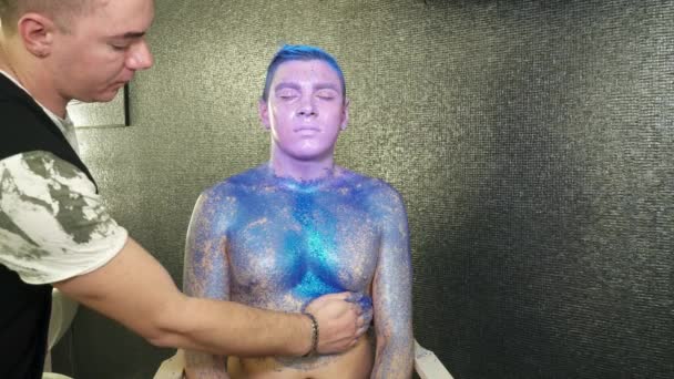 Митець тіла, що покриває тіло молодого чоловіка блакитними блискітками — стокове відео