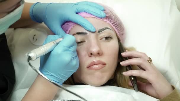 Cosmetologist is making an eyebrow permanent makeup while woman talking on the phone — Stock Video