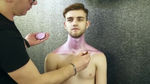 Художник-макіяж, що покриває шию молодого чоловіка фарбою — стокове відео