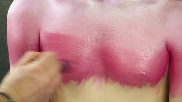 Artista de maquillaje cubriendo el pecho de los hombres jóvenes con una pintura rosa — Vídeos de Stock