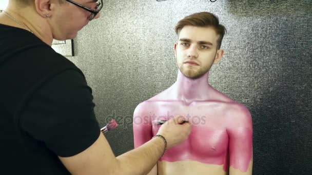 Визажист, покрывающий тело молодого человека розовой краской в студии — стоковое видео