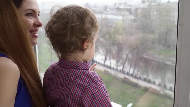 Молодая красивая мать смотрит в окно со своим маленьким мальчиком замедленной съемки — стоковое видео