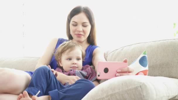 年轻的母亲和她的儿子坐在沙发上看动画片对智能手机 — 图库视频影像