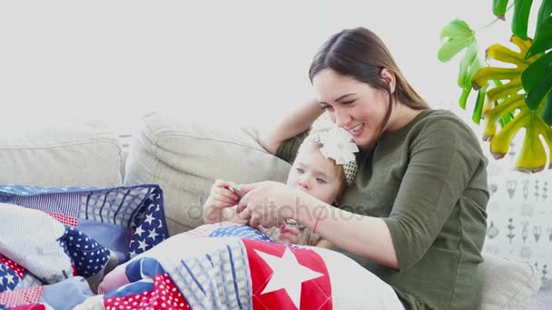 Madre usando el celular con su hija acostada en el sofá debajo de la manta en la habitación — Vídeo de stock