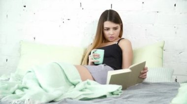 Yatakta oturmuş ve bir fincan bir kitap okuma güzel kadın