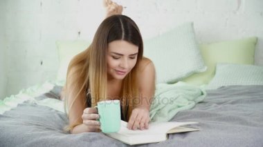 Bir fincan ile yatağın üstüne oturan ve bir kitap okuma zevk güzel kadın