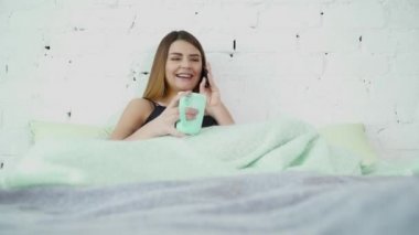 Genç kadın laughng ve yatakta battaniyenin altında oturan ve telefonda konuşurken