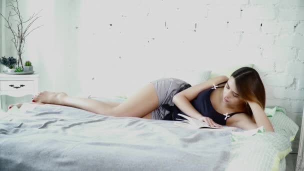 Молодая женщина в пижаме лежит на кровати и читает книгу — стоковое видео