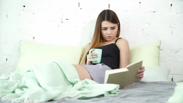 Gyönyörű nő ült az ágyon, és olvasott egy csésze — Stock videók