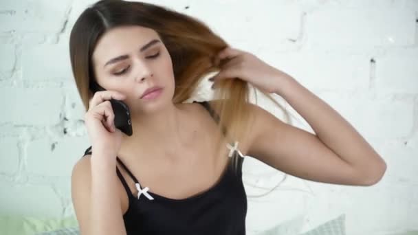 Unga kvinnan sitter på sängen under en filt leende och pratar i telefon — Stockvideo