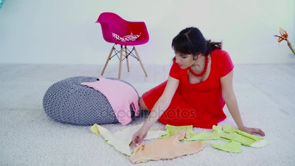 Беременная женщина сидит на ковре и выбирает одежду для будущего ребенка — стоковое видео