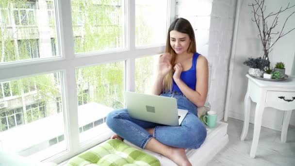 Молода жінка за допомогою ноутбука говорить на скайпі, сидячи на підвіконні — стокове відео