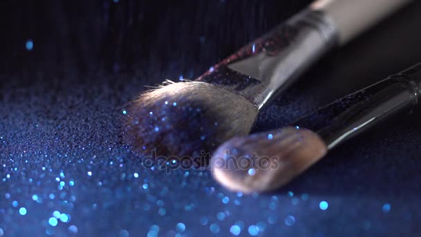 Brillos brillantes azules que caen sobre cepillos de maquillaje en el fondo negro, cámara lenta abstracta — Vídeo de stock