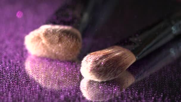 Paillettes violettes tombant sur les pinceaux de maquillage sur le miroir sur le fond noir, au ralenti — Video