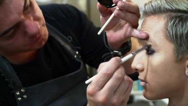 Artista de maquillaje masculino haciendo arte facial para un joven hombre rubio — Vídeos de Stock