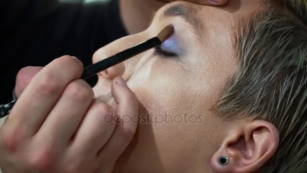 Maquillaje artista haciendo arte facial para un joven hombre rubio, pintando los ojos de cerca — Vídeo de stock