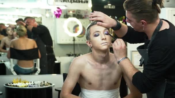 Maquillaje artista haciendo arte facial para un joven hombre rubio delante del espejo — Vídeos de Stock
