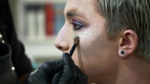 Maquillaje artista haciendo arte facial con destellos para un hombre rubio con un primer cepillo de maquillaje — Vídeos de Stock