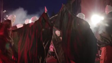 Ayaklık takan dansçılar pelerin giyer, el kaldırır, tuhaf görünürler..