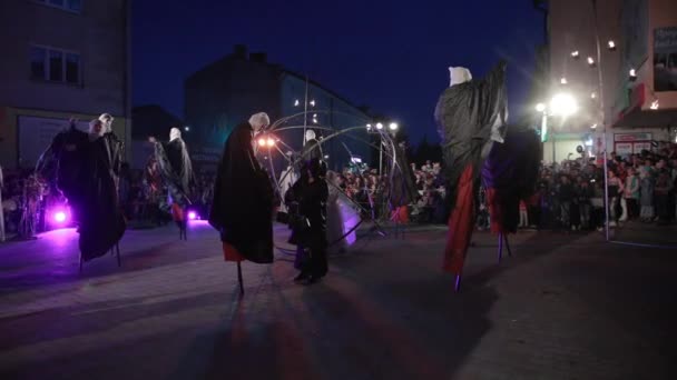 Man in zwart deelt kommen uit aan steltlopers, vrouw met lantaarn in bol. — Stockvideo