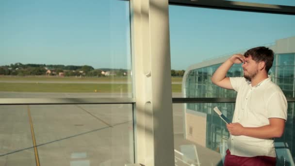 Человек с бородой, стоит в аэропорту у окна, держит паспорт, ждет самолет — стоковое видео