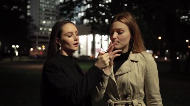 Jonge mooie vrouw helpt meisje om een sigaret aan te steken. Nacht stad op de achtergrond. — Stockvideo