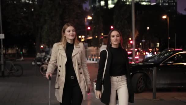 Két csinos lány gurítja a bőröndjét az éjszakai városban.. — Stock videók