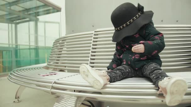 Шляпа падает на красную голову маленькой девочки, сидящей на скамейке с кулинаром — стоковое видео