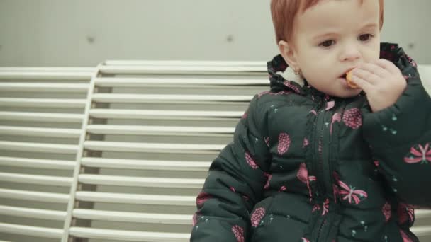 Маленькая рыжеволосая девочка ест печенье на белой скамейке — стоковое видео