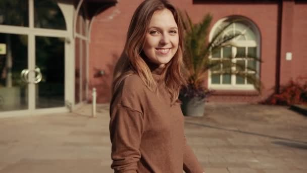 Жінка коричневе волосся коричневий светр йде від камери, крутиться навколо, посміхається — стокове відео