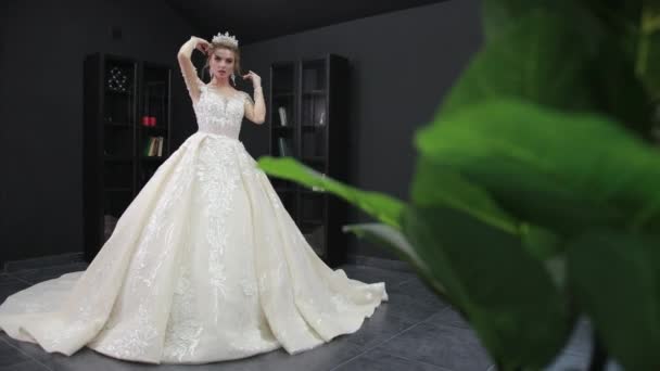 Mladá blondýnka nevěsta v bílých svatebních šatech s korunou představuje pro kameru v pokoji — Stock video