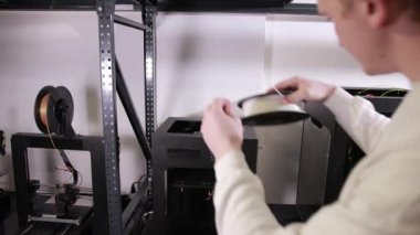 3D technology young man inserts red plastic thread cable from coil into printer