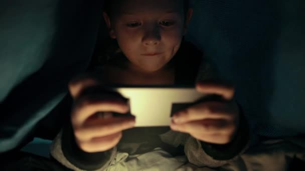 Little guy under the blanket is playing a game on his phone smartphone at night — Stockvideo