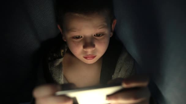 Little guy under the blanket is playing a game on his phone smartphone at night — Wideo stockowe